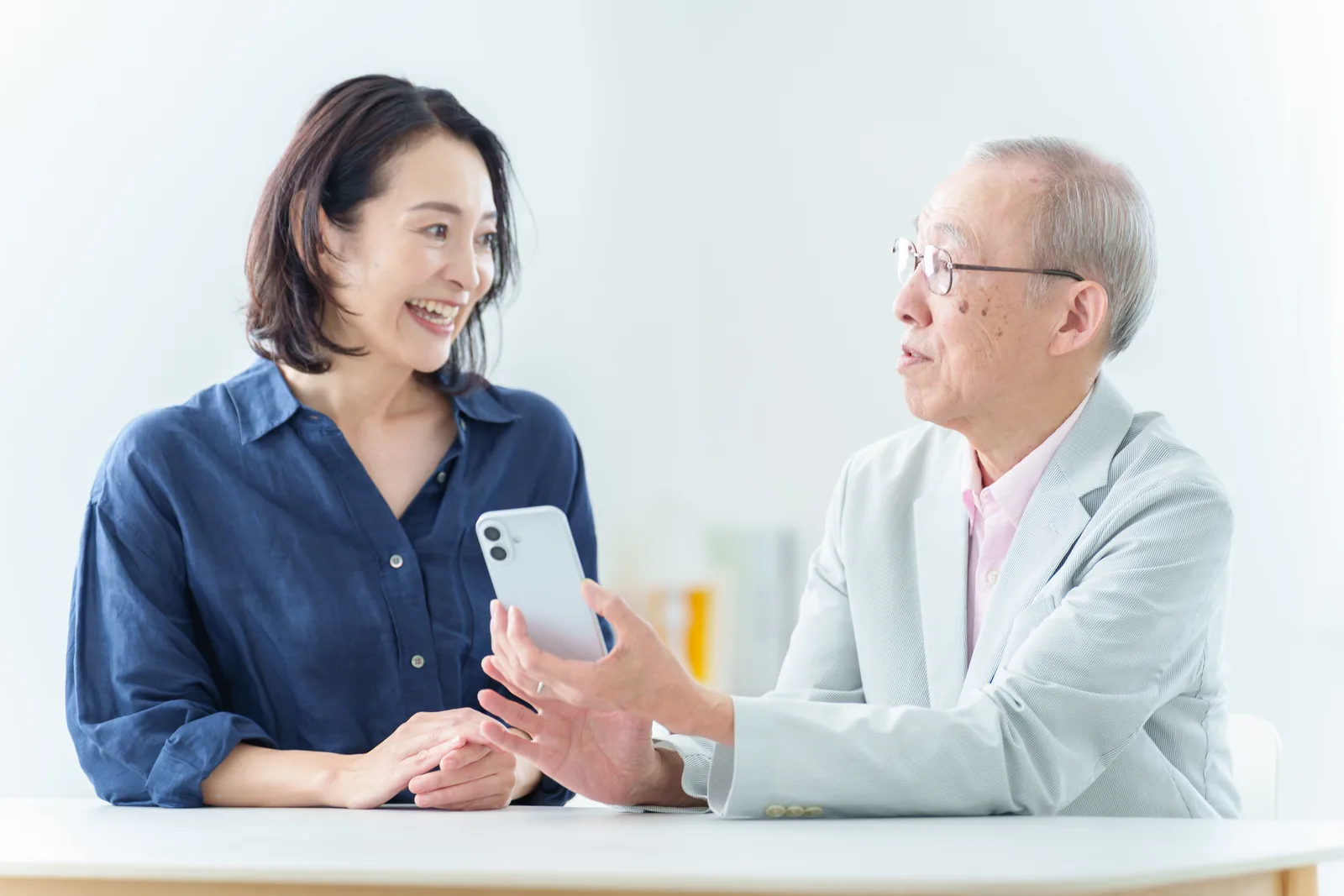明るい室内の白いテーブルで、娘さんと白髪のお父さまが少し離れて並んで座り、お父さまが手に持ったスマートフォンを二人の間に掲げ、娘さんが笑ってお父さまのほうを見ている様子