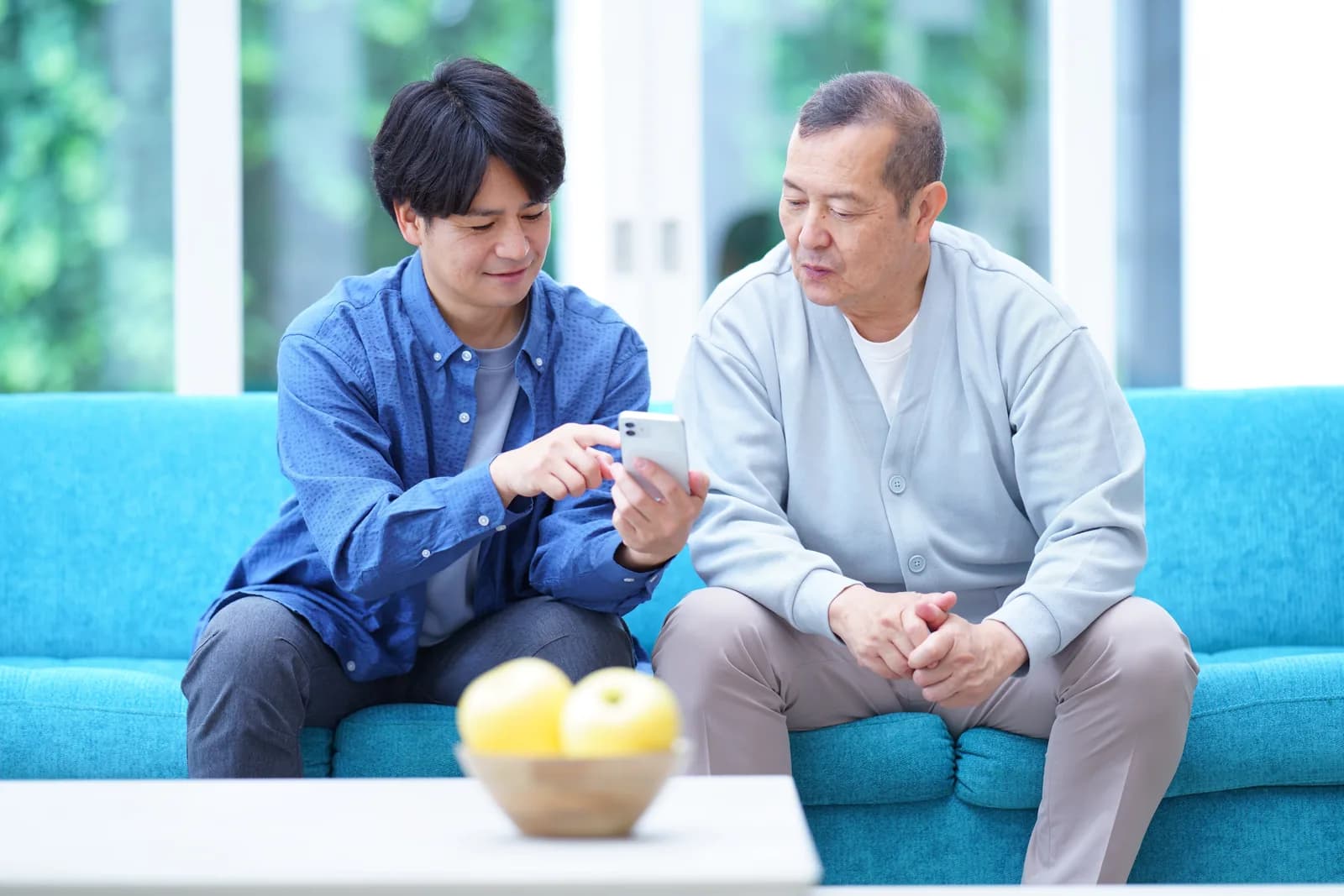 青いソファに並んで座った息子さんが、お父さまにスマートフォンの画面を指さしながら見せている様子