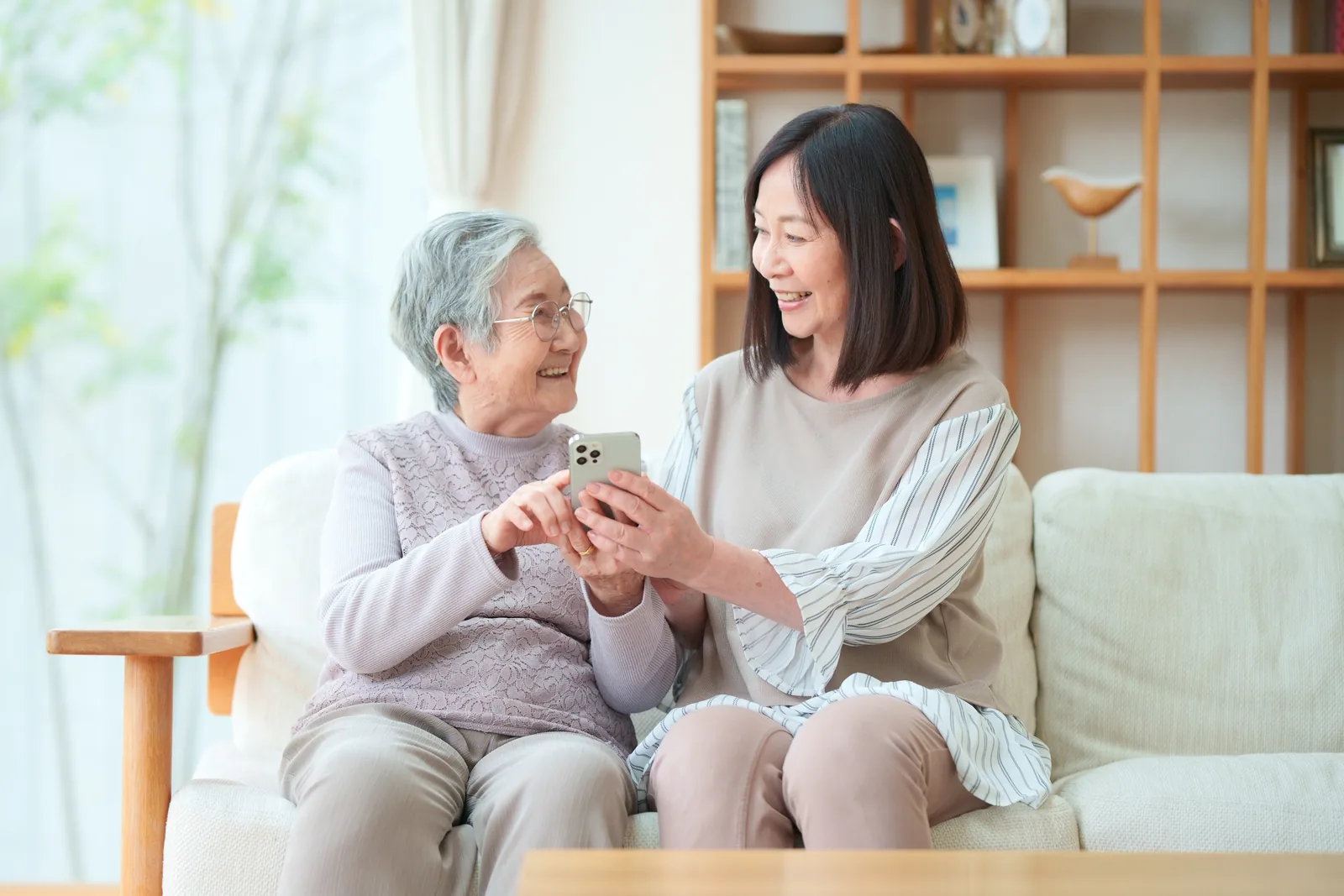 明るい居間のソファで、白髪のお母さまと娘さんが並んで座り、一緒にスマートフォンの画面を見て笑っている様子